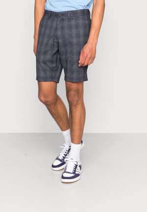 Dunkle Karoshorts aus leichtem Stoff mit Knopfverschluss und Seitentaschen. Kombiniert mit weißen Socken sowie marineblauen und weißen Sneakern.