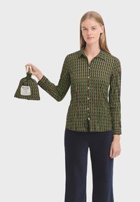 Groen en bruin gestreept shirt met verticale zigzaglijnen, lange mouwen en een klassieke kraag. Houdt een bijpassende tas vast.