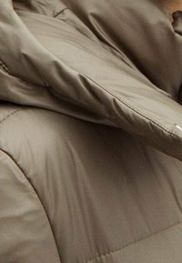 Veste matelassée en taupe doux, dotée d'un col montant et d'une texture lisse, avec des coutures apparentes et un pli au niveau de l'encolure.