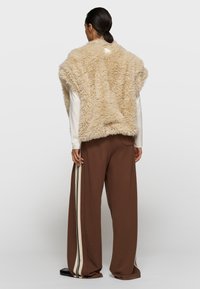 Chaleco de piel beige con textura esponjosa, usado sobre una camiseta blanca de manga larga y pantalones marrones de pierna ancha con una franja blanca a lo largo del costado.