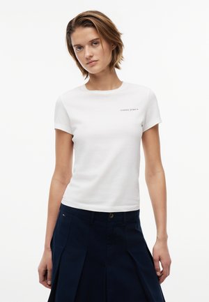 Νεαρή γυναίκα που φοράει λευκό μπλουζάκι Tommy Jeans και σκούρα μπλε πλισέ φούστα, στέκεται μπροστά σε απλό λευκό φόντο.