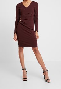 Burgundy långärmad klänning med V-ringning, samlad nederdel fram och tre guldfärgad knappdetaljer på sidan. Tillverkad av en slät och mjuk tyg.