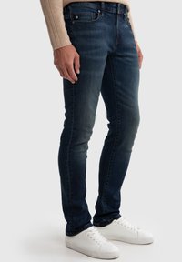Jeans de denim azul oscuro con corte ajustado, que presentan un sutil desvanecimiento, diseño clásico de cinco bolsillos y un pequeño parche de marca en la cinturilla.