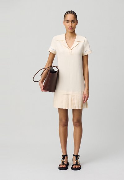 Vestido corto de crema con cuello tipo camisa y falda plisada. La modelo sostiene un bolso tote marrón. Las sandalias negras realzan el conjunto.