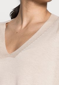 Närbild av en person som bär en beige V-ringad stickad tröja, som visar halsen, nyckelbenet och övre bröstområdet.