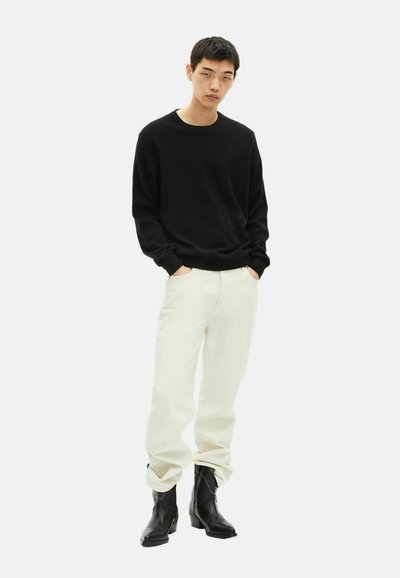 Młody mężczyzna ubrany w czarny sweter z długim rękawem, jasnobeżowe spodnie i czarne skórzane buty, stojący z rękami w kieszeniach.