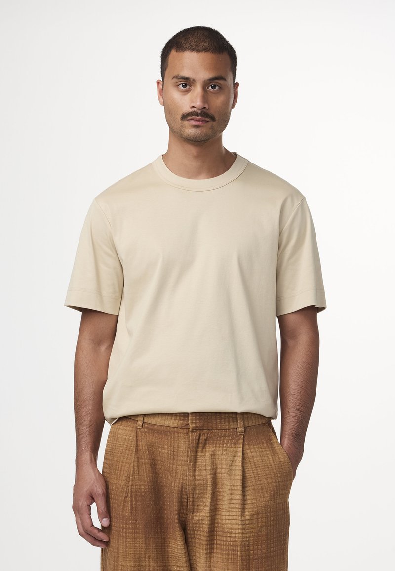 T-shirt beige a maniche corte con scollatura rotonda, realizzato in tessuto morbido. Indossato con pantaloni leggeri marrone chiaro texture a griglia.