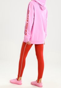 Felpa rosa con cappuccio e tasca frontale, caratterizzata da testo bianco in grassetto; abbinata a leggings rossi con strisce bianche ai lati e sneakers rosa.