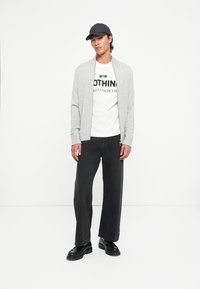 Grijze cardigan, wit t-shirt met zwarte tekst, zwarte wijde jeans en zwarte schoenen; model draagt een zwarte pet. Eenvoudig ontwerp, casual pasvorm.