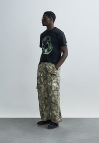 Μαύρο μπλουζάκι με πράσινο γραφικό σχέδιο, σε συνδυασμό με παντελόνι καμουφλάζ cargo. Μαλακή υφή, φαρδιά εφαρμογή, φορεμένο με μαύρα slip-on παπούτσια και γυαλιά ηλίου.