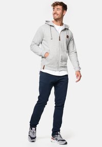 Svijetlosivi zip-up dukserica s podesivim kapuljačom, dva bočna džepa i smešnim kožnim detaljima. Nosena preko bijele majice i uparena s mornarskim hlačama.
