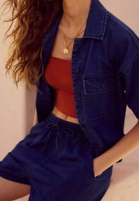 Granatowa koszula dżinsowa z krótkimi rękawami, zestawiona z dopasowanym, rdzawym topem oraz dopasowanymi granatowymi szortami. Srebrne, złote naszyjniki na warstwy ozdabiają dekolt.