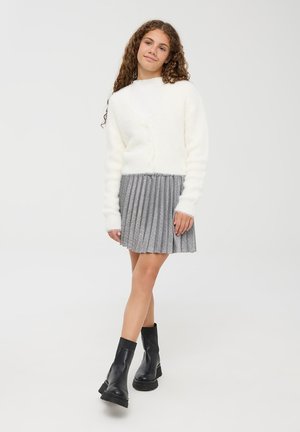 Cardigan bianco e soffice sopra una maglietta chiara, abbinato a una gonnellina grigia plissettata con una leggera lucentezza. Completato con stivali neri alla caviglia.
