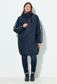 Parka azul marino impermeable con capucha, que presenta rayas plateadas en las mangas, dobladillo elástico y bolsillos frontales con cremallera. Textura suave.