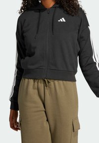 Svart zip-up hoodie med huva, vita strimmor längs ärmarna, och en liten vit logotyp. Matchat med olivgrön byxor med elastisk midja.