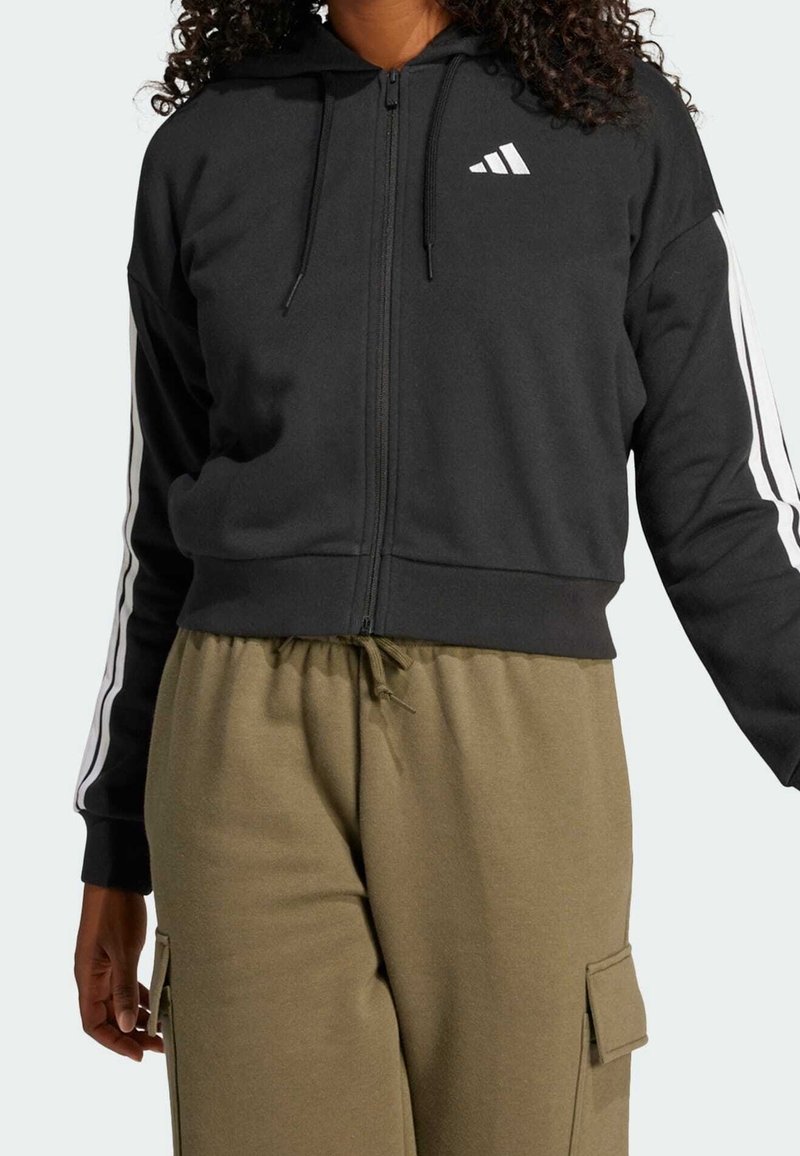 Svart zip-up hoodie med huva, vita strimmor längs ärmarna, och en liten vit logotyp. Matchat med olivgrön byxor med elastisk midja.