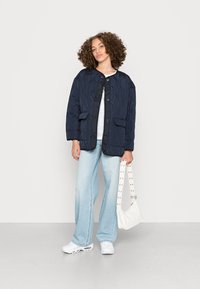 Giacca imbottita blu navy con grandi tasche, caratterizzata da un colletto rotondo e bottoni frontali. Il modello indossa jeans svasati azzurri chiari e scarpe da ginnastica bianche.