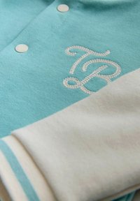 Turkusowa koszula z białymi akcentami, z haftowanymi inicjałami. Jasne guziki i podwinięte rękawy z detalem w paski.