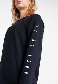 Sweatshirt noir en tissu doux. Il présente un col rond et des manches longues avec l'inscription « JUST DO IT » imprimée en blanc le long de la manche droite.