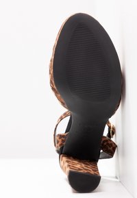 Sandale femme en imprimé léopard avec un talon bloc, une bride cheville et une semelle texturée noire, vue par en dessous sur fond blanc.