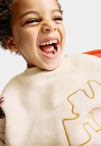 Beige tröja med ribbad krage, prydd med en guldbroderad design. Barn med lockigt hår som skrattar brett och visar tänderna.