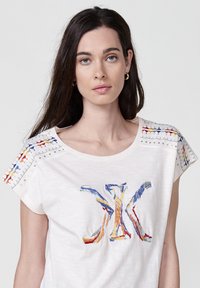 Chemise blanche à manches courtes avec broderies multicolores sur les épaules et à l'avant. Présente des coutures décoratives et des accents métalliques.