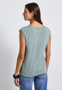 Jasnozielony bezrękawnik z okrągłym dekoltem, gładką fakturą i przeszytymi krawędziami, noszony z niebieskimi dżinsami.