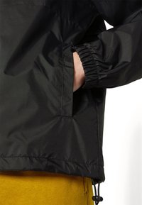 Ręka w czarnej kurtce z elastycznym mankietem i sznurkiem w dolnej części, pod kurtką widoczne musztardowożółte spodnie.