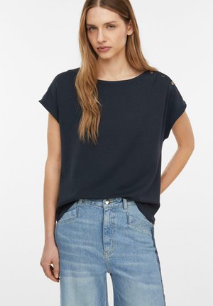 Kobieta o długich jasnobrązowych włosach, ubrana w granatowy krótki rękaw z guzikami na ramionach oraz jasnoniebieskie wysokie spodnie dżinsowe.