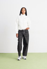 Balta zip-up jaka ar Nike logo, melnas rakstainas platās bikses un pelēki sporta apavi. Vienkārša fona ar zaļu grīdu.