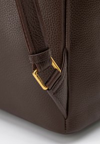 Sac à main en cuir marron avec une finition texturée. Présente un clip en métal doré et une sangle lisse. Forme carrée avec des lignes angulaires.