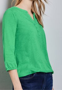 Grön blus med långa ärmar och henley-krage, tre knappar och muddade ärmar. Tillverkad av lätt, strukturerad tyg.