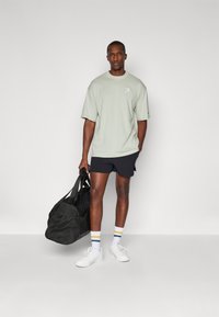 Lysgrøn oversized T-shirt med logo, sorte shorts, hvide sneakers og flerfarvede stribede sokker. Bærer en sort duffel-taske.