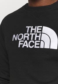 Svart sweatshirt med rund halsringning, med en framträdande vit broderad logotyp av "THE NORTH FACE" och en böjd designelement.