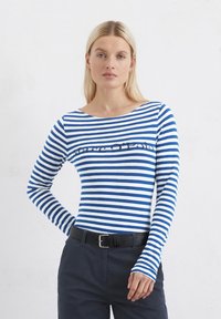 Haut rayé à manches longues en coton bleu et blanc avec un col bateau et un logo imprimé sur la poitrine, associé à un pantalon sombre et une ceinture noire.