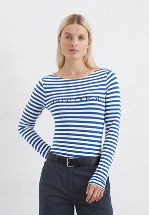 Gestreiftes langärmliges Oberteil in Blau und Weiß aus Baumwolle mit einem Bootsausschnitt und einem aufgedruckten Logo auf der Brust, kombiniert mit dunklen Hosen und einem schwarzen Gürtel.