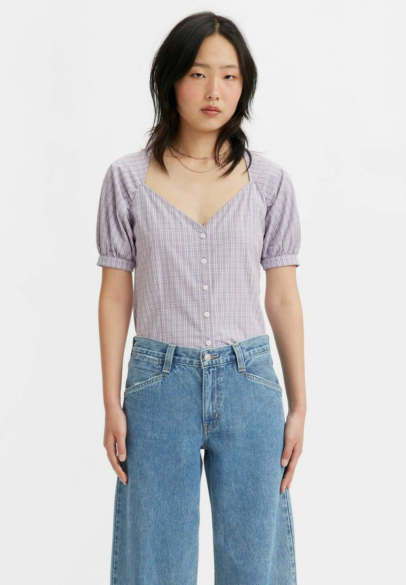 Blusa a quadri viola con maniche a sbuffo, scollo a V e chiusura frontale con bottone, abbinata a jeans in denim a gamba larga blu chiaro.