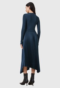 Kaksi sininen ribbed-mekko pitkillä hihoilla, jossa on laskostettu hame ja korkealaatuinen helma, yhdistettynä mustiin nilkkureihin.