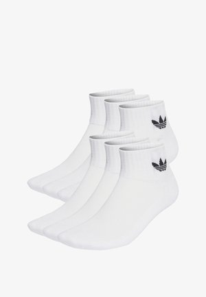 Calzini bianchi in cotone fino alla caviglia con polsini a coste e logo Adidas nero. Cinque paia disposti in fila, mostrando una texture liscia e una forma uniforme.