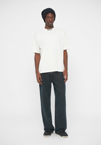 Λευκό κοντομάνικο μπλουζάκι με έντονη γραφή, σε συνδυασμό με σκούρες φαρδιές τζιν παντελόνες και μαύρα αθλητικά παπούτσια, συμπληρωμένο με ένα μαύρο πλεκτό καπέλο.