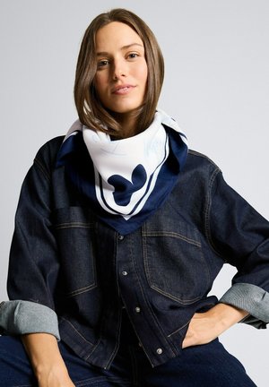 Jeune femme aux cheveux bruns portant une écharpe blanche et marine ainsi qu'une veste en jean foncé, posant avec la main sur la hanche devant un fond uni.