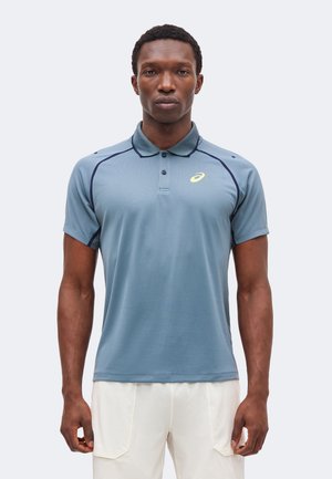 Muž nosiaci modré polo tričko s krátkym rukávom so žltým logom Asics a biele nohavice, stojaci na jednolícom pozadí.