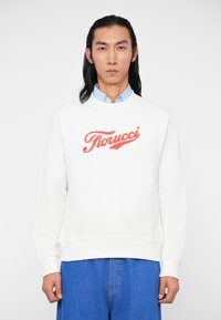 Sweatshirt branco com decote redondo e punhos canelados, apresentando um ousado logotipo vermelho "Fiorucci" atravessado no peito. Combinado com calças de ganga azuis.