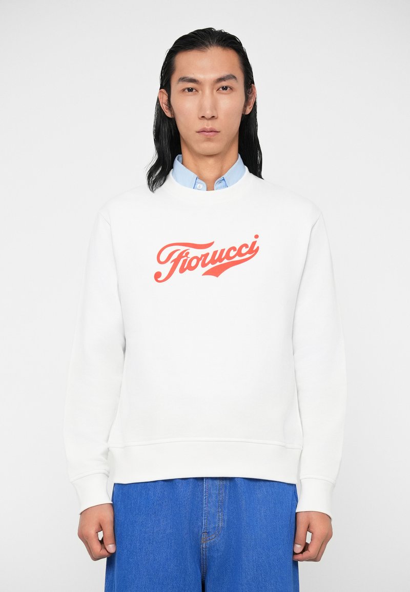 Sweatshirt branco com decote redondo e punhos canelados, apresentando um ousado logotipo vermelho "Fiorucci" atravessado no peito. Combinado com calças de ganga azuis.