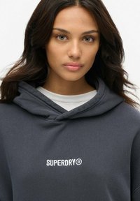 Sudadera con capucha de color gris oscuro con un logo blanco de "SUPERDRY" en el pecho, con una textura suave y un corte relajado.