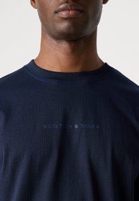 Kleita tumši zila kokvilnas T-kreklā ar apaļu kaklu un izsmalcinātu priekšējo druku ar zīmola nosaukumu "SCOTCH & SODA" gaišākā tonī.