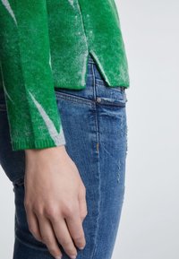 Un pull en laine vert avec une texture douce, orné de motifs abstraits blancs. Assorti à un jean en denim léger et déchiré bleu.