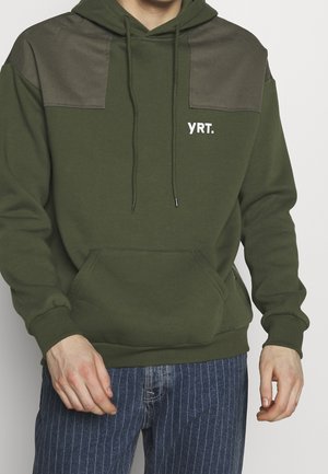 Man draagt een olijfgroene hoodie met donkerdere schouderstukken en witte "YRT."-tekst, gecombineerd met blauwe spijkerbroek met krijtstreepjes, handen ontspannen langs zijn zij.