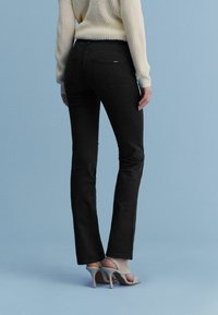 Schwarze Denim-Jeans mit weit ausschwingendem Bein und glatter Textur, mit Gesäßtaschen und mittelhohem Bund, kombiniert mit hellen Schuhen.