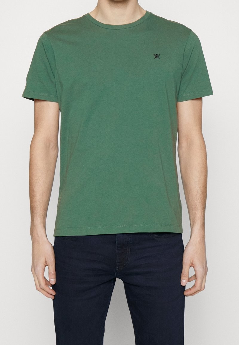 T-shirt en coton vert avec un col rond, des manches courtes et un logo noir discret sur le côté gauche de la poitrine. Texture lisse et coupe classique.